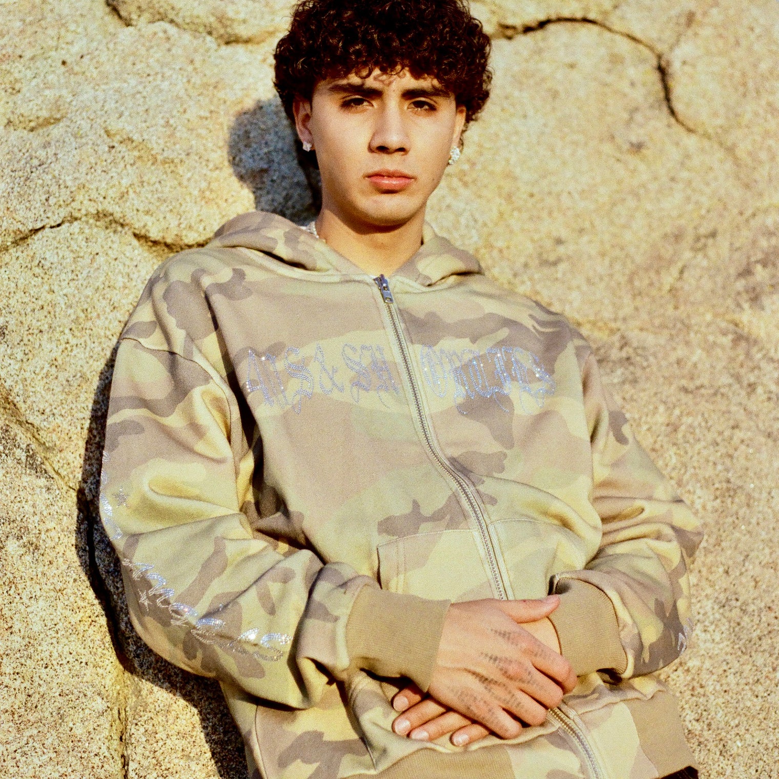 Model wearing 40s & Shorties Angel Wings Hoodie in camo with rhinestone logo across chest, standing against a rock wall.