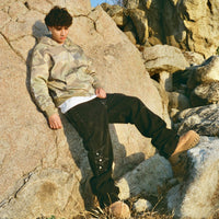 Model in 40s & Shorties Angel Wings Hoodie camo print with rhinestone graphics, styled with black pants and boots, seated on boulders outdoors.