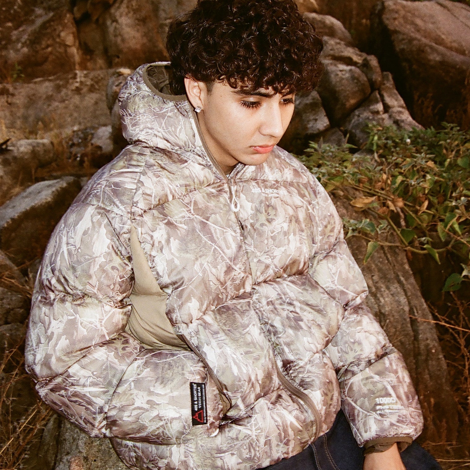 Model wearing 40s & Shorties Faded Camo Puffer Jacket with tonal camouflage print and hood, sitting outdoors on rocky terrain.