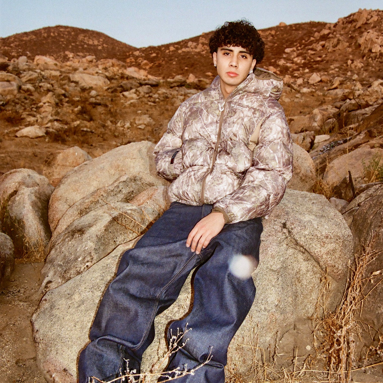 Model in 40s & Shorties Faded Camo Puffer Jacket with hood, styled with wide-leg denim, seated on rocks in desert landscape.