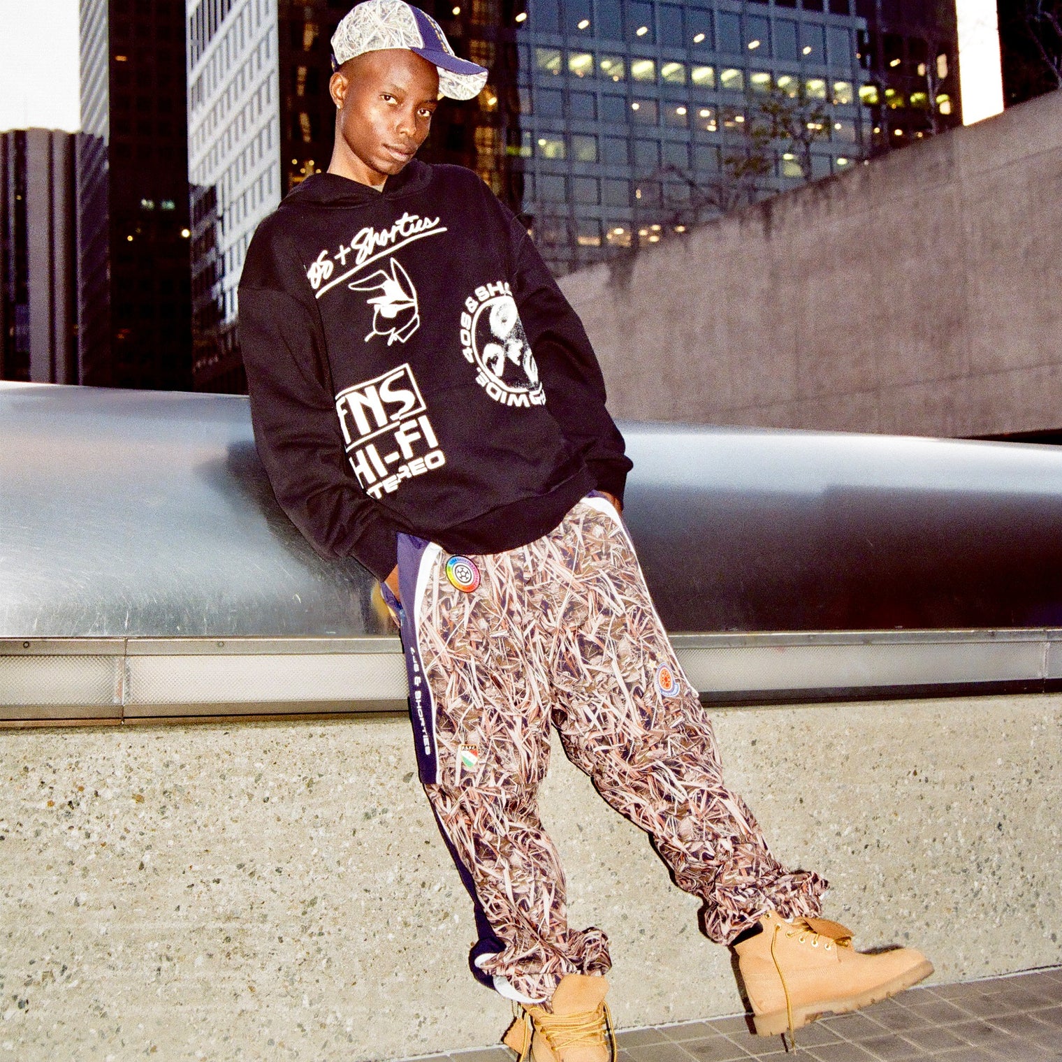 Model wearing the Layout Hoodie on the street styled with patterned pants and boots, city skyline in the background.