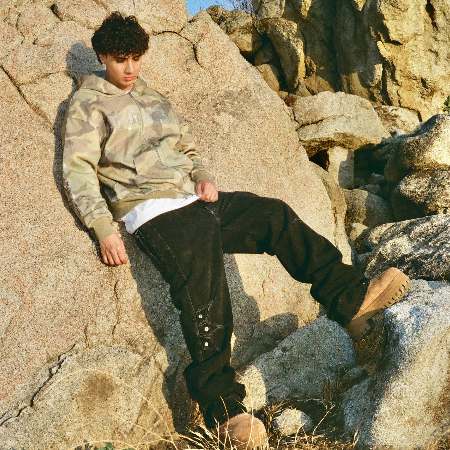 Model in 40s & Shorties Angel Wings Hoodie camo print with rhinestone graphics, styled with black pants and boots, seated on boulders outdoors.

