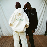 Models facing away wearing French General Zip Hoodies in cream and black, showing bold back text prints – 40s & Shorties.
