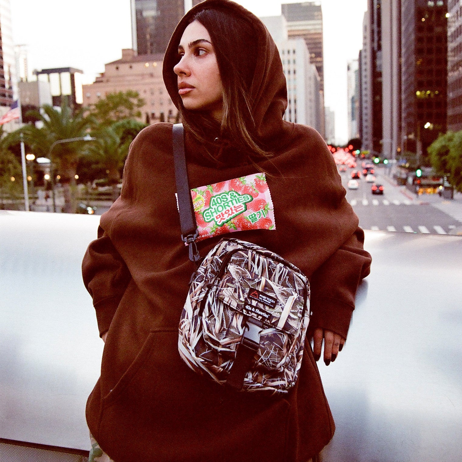 Woman wearing brown Gummy Hoodie and camo crossbody bag on a city street bridge
