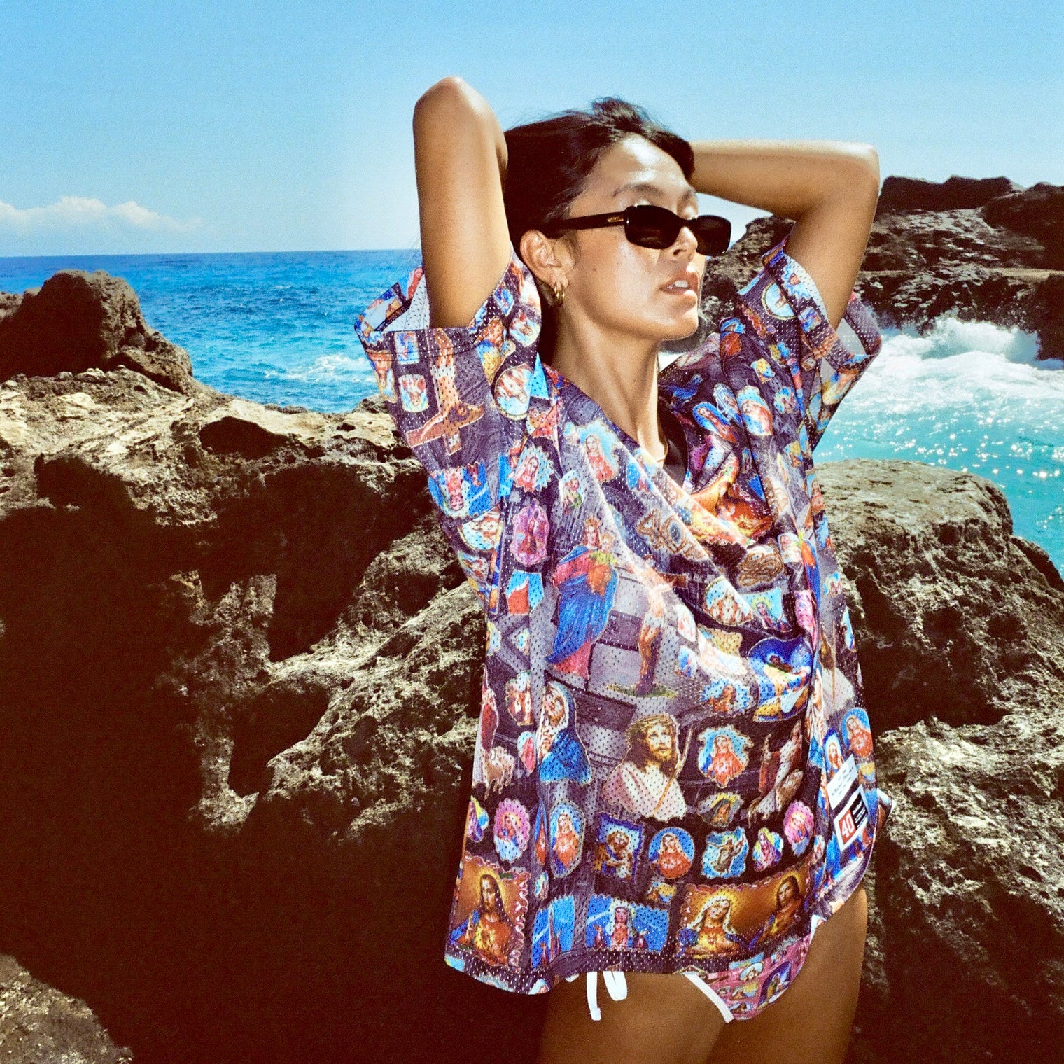 Female model stretching arms behind head while wearing Holy Spirit Football Jersey over bikini, by ocean rocks