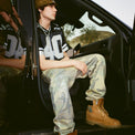 Model wearing 40s & Shorties Rhinestone Sweatpants in camo with rhinestone logo detail, paired with a black Fubar Jersey, seated in a car doorway.