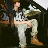 Model wearing 40s & Shorties Rhinestone Sweatpants in camo with rhinestone logo detail, paired with a black Fubar Jersey, seated in a car doorway.