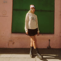 Female model standing in sunlight wearing the Scribble Logo Sweater in Cream with boots and a skirt