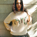 Model in Scribble Logo Sweater in Cream posing with hands on hips against a concrete wall