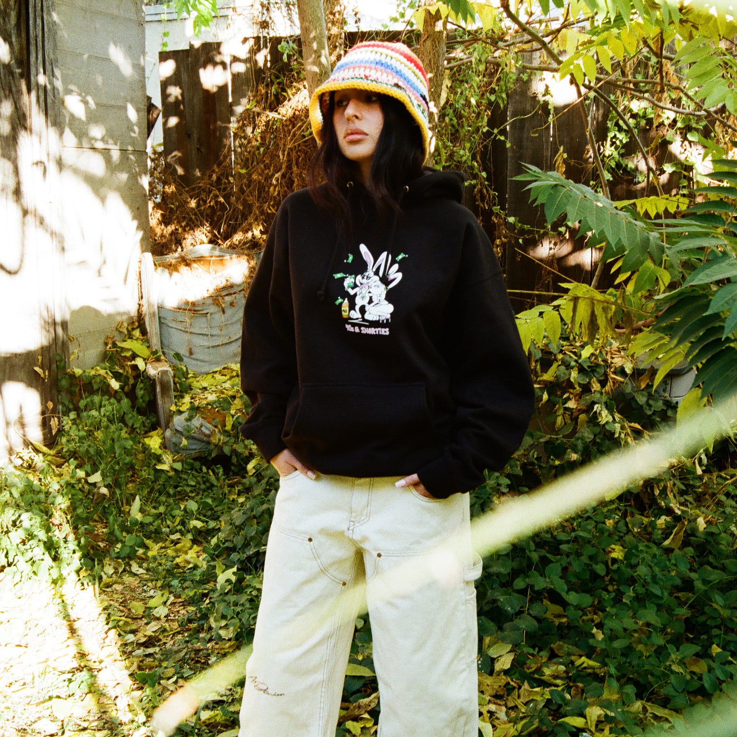 Model wearing black 40s & Shorties hoodie with front graphic, standing outdoors in a leafy setting.
