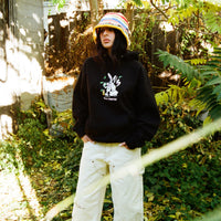 Model wearing black 40s & Shorties hoodie with front graphic, standing outdoors in a leafy setting.
